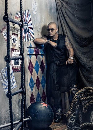 Stylish bald man with tattoos leans against a patterned surface in a moody indoor setting with ropes and vintage posters.