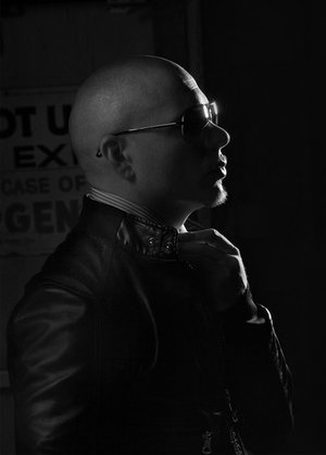 Monochrome portrait of a bald adult male in sunglasses and leather jacket adjusting his collar in a dimly lit indoor setting.