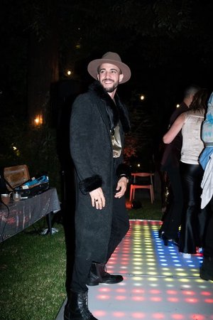 Adult male in a black coat and hat posing on a colorful dance floor at night, with people and tables in the background.