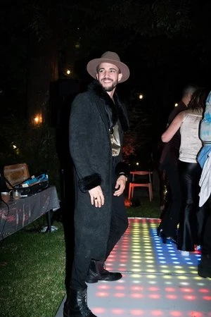 Adult male in a black coat and hat posing on a colorful dance floor at night, with people and tables in the background.