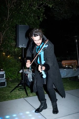 Adult male plays a blue electric violin outdoors at night, wearing a black coat and boots, with a speaker and other equipment visible.
