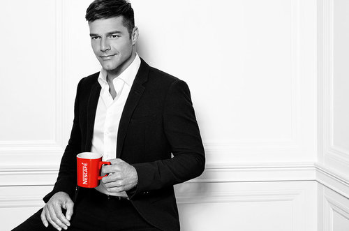 Handsome man in a suit smiles while holding a bright red mug against a minimalist white wall, creating a striking portrait.