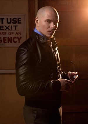 Portrait of a bald adult male wearing a black jacket and holding glasses, posing in an indoor setting with an emergency exit sign visible.