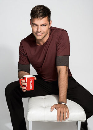 Adult man seated, holding a red Nescafé mug, in a studio setting with a white backdrop and stool. He is wearing a maroon shirt and black pants.