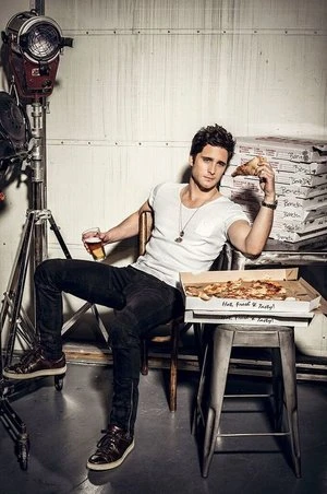 Handsome man relaxes in a studio with pizza and a beer, casually seated with a soft light backdrop, creating a stylish editorial feel.