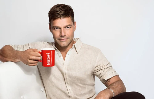 Adult male celebrity poses indoors in an eye-level medium shot, holding a red Nescafé mug, and wearing a beige linen shirt.