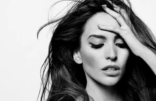 Black and white portrait of a woman with long, dark hair holding her hand to her forehead, eyes closed, against a light backdrop.