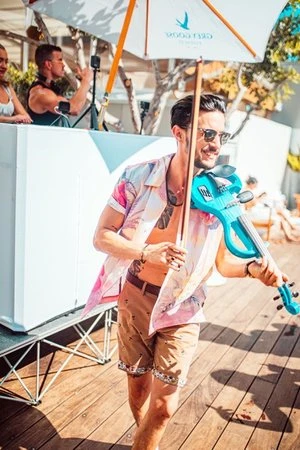 Man in a floral shirt playing a blue electric violin on a wooden deck with a Grey Goose umbrella and DJ in the background.