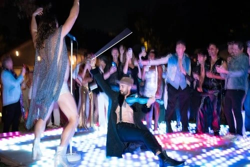 Two performers entertain a crowd on a lit stage at night, with one playing violin and the other dancing in a sparkling outfit.