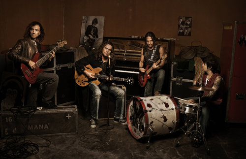 Four musicians playing instruments in a dimly lit music studio with guitars, a drum set, and a piano in a band performance.