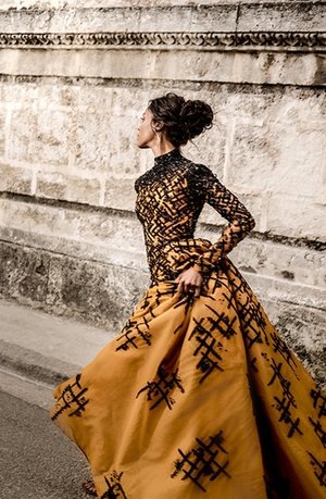A woman in a long gold dress with black embellishments poses in front of a stone wall, styled with a high bun and looking elegant.
