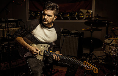 Adult male musician playing a black electric guitar in a music studio setting with visible amplifier, drum set, and shelf of awards.