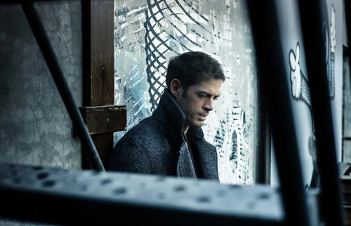 Man in dark coat seated against graffiti wall. Moody, introspective portrait. Urban setting. Focus on person, weathered background.