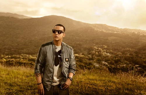 A man in sunglasses stands on a grassy hill against a backdrop of forested mountains under a hazy, bright sky. He wears a jacket and jeans.