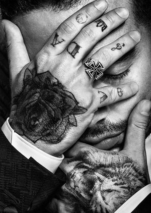 Close-up black and white studio portrait of a man's tattooed hands covering his face, wearing a ring and suit jacket.
