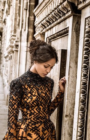A woman with an updo hairstyle wearing an ornate patterned dress standing next to a carved stone wall.