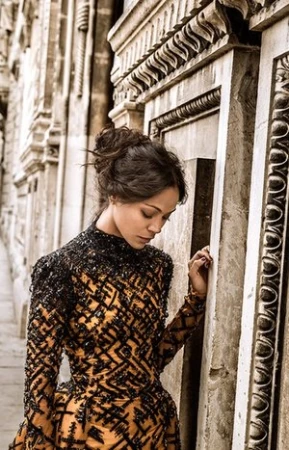 A woman with an updo hairstyle wearing an ornate patterned dress standing next to a carved stone wall.