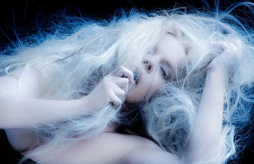 Close-up portrait of musician Maury Hechavarria with wild white hair framing her face against a dark background