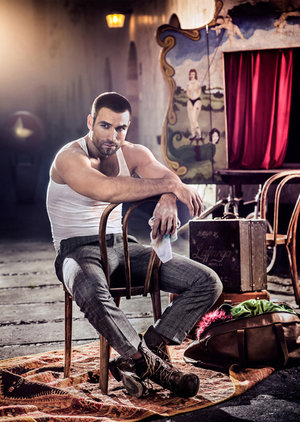 Muscular man in tank top and grey trousers poses in front of a vintage circus backdrop, seated on chair, vintage luggage beside him.