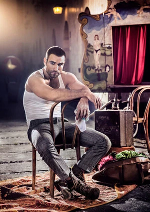 Muscular man in tank top and grey trousers poses in front of a vintage circus backdrop, seated on chair, vintage luggage beside him.