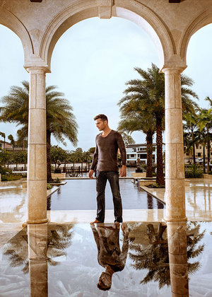 Man stands in arched walkway with reflection in water, framed by palm trees and light stone pillars, creating a serene architectural portrait.