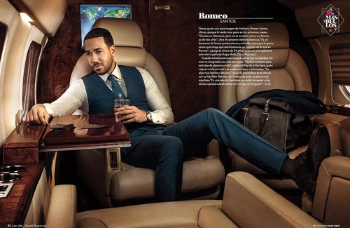 Handsome man in teal suit and vest sits in private jet with drink. Polished wood table, Louis Vuitton bag, airplane window. Editorial portrait.