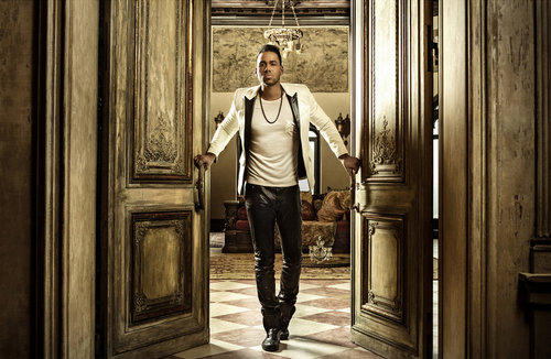 Portrait of an adult male standing in a doorway wearing a white jacket, black pants, and black shoes inside an ornate room.