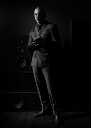 Bald man in dark suit stands near speakers in dim light. Low-key portrait with emphasis on texture and sharp tailoring in monochrome.