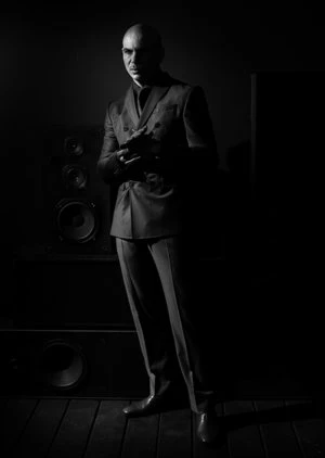 Bald man in dark suit stands near speakers in dim light. Low-key portrait with emphasis on texture and sharp tailoring in monochrome.