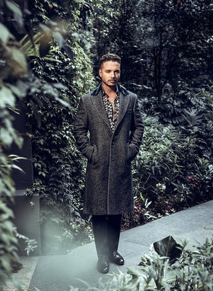 Adult male posing in a patterned shirt and grey overcoat in front of green foliage, creating a sophisticated and stylish outdoor portrait.