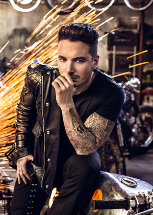 Portrait of adult male with tattoos in black shirt and jacket sitting on a motorcycle inside with sparks flying in the background.