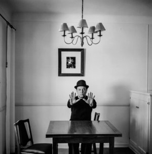 A person in a bowler hat sits at a table with a raised hand, a framed photo on the wall, lamps, and chairs in a monochrome setting.