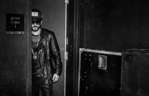 Black and white portrait of a man standing in a doorway wearing a leather jacket, sunglasses, and a baseball cap inside a green room.