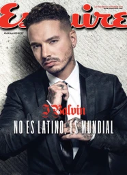 Portrait of J Balvin wearing a black suit and white shirt, holding his tie against a textured stone wall.