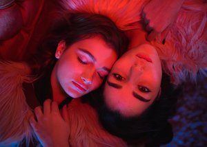 Close-up studio portrait of two young adult women, lying head-to-head. One woman has dark hair; the other has lighter hair, both fair skin.