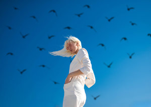 An adult female stands against a bright blue sky as a flock of dark birds flies around her, creating a striking contrast.