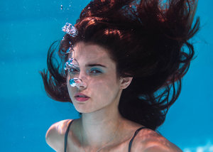 Underwater portrait of a woman with brown hair, partially submerged, in blue-toned water with visible air bubbles around her face.