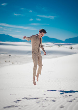 A young adult male in a tan jumpsuit leaps barefooted in a white desert landscape with a clear blue sky and distant mountains.