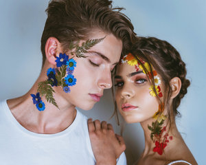 A young couple poses in a close-up studio portrait featuring floral face art against a blue background.
