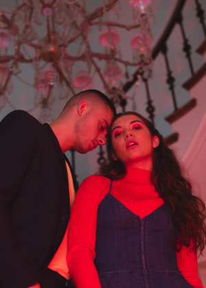Man and woman in studio, bathed in red light from above chandelier. Woman in denim dress and red turtleneck, man in black jacket.
