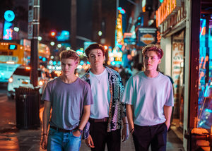 Three young men stand on a city sidewalk at night under bright neon lights from businesses. The scene has a vibrant, youthful feel.