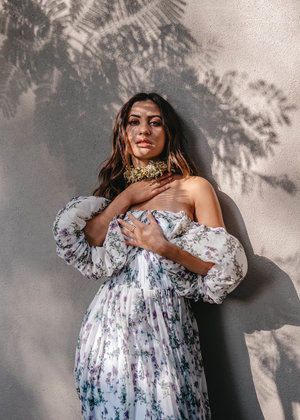 Woman in floral dress posing against a wall with shadow patterns, wearing a floral collar. Elegant outdoor portrait.