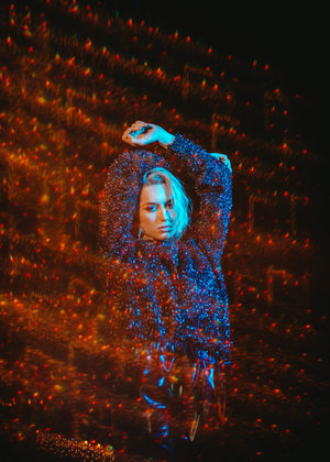 Portrait of a woman in a sparkling top, arms raised against a black background with bokeh lights in blue and red, medium shot.