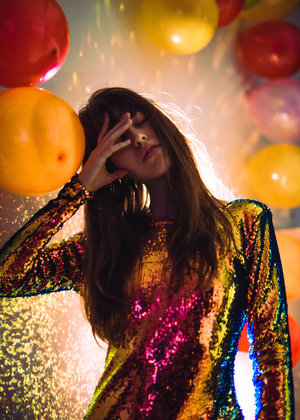 Woman with long brown hair in sequin dress touches forehead standing indoors with balloons on wall in soft, diffused lighting.