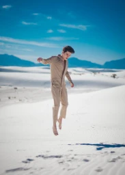 A young man in a beige jumpsuit mid-air jumping over white sand dunes with blue mountains and sky behind him.