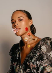 Portrait of a Black woman holding a pink flower in her mouth with colorful pressed flowers on her neck and wearing a floral jacket