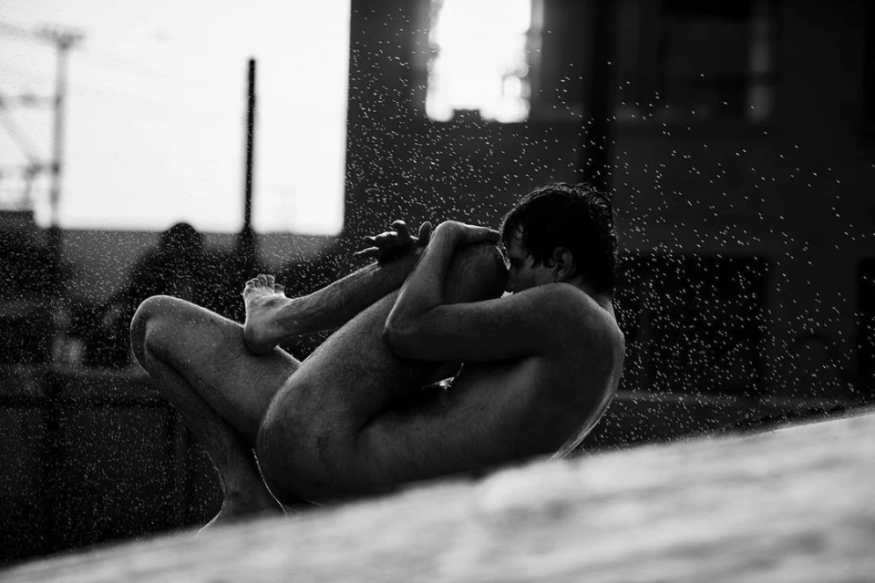 Monochrome image of a nude adult man in a seated yoga position outdoors, with water droplets in the air and an urban backdrop.