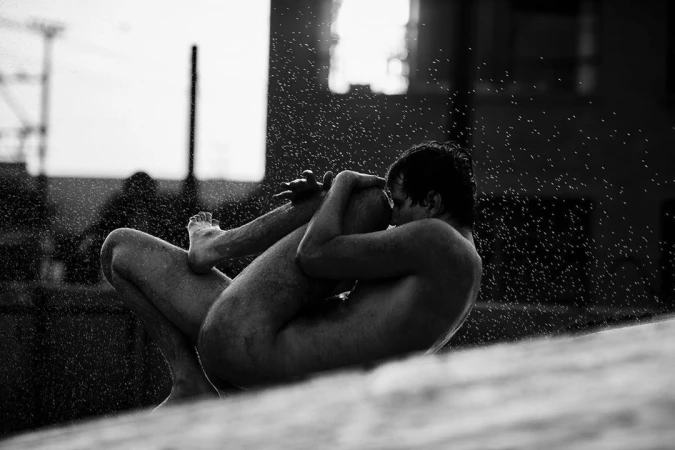 Monochrome image of a nude adult man in a seated yoga position outdoors, with water droplets in the air and an urban backdrop.
