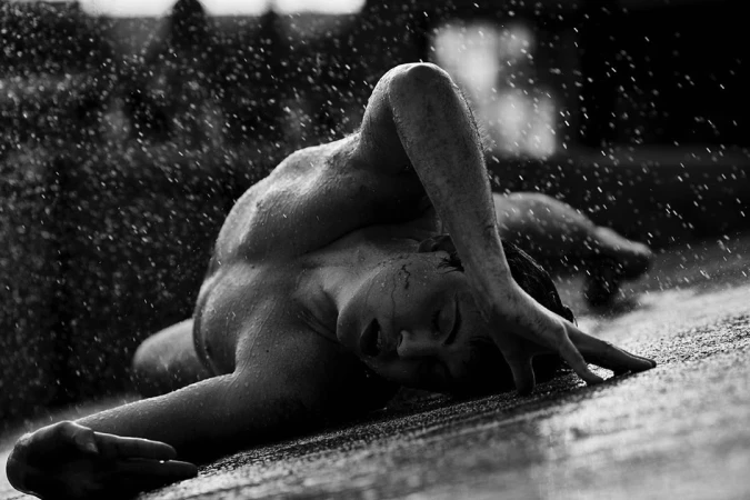 Monochrome low-angle shot of a person lying on wet ground surrounded by heavy falling water drops.