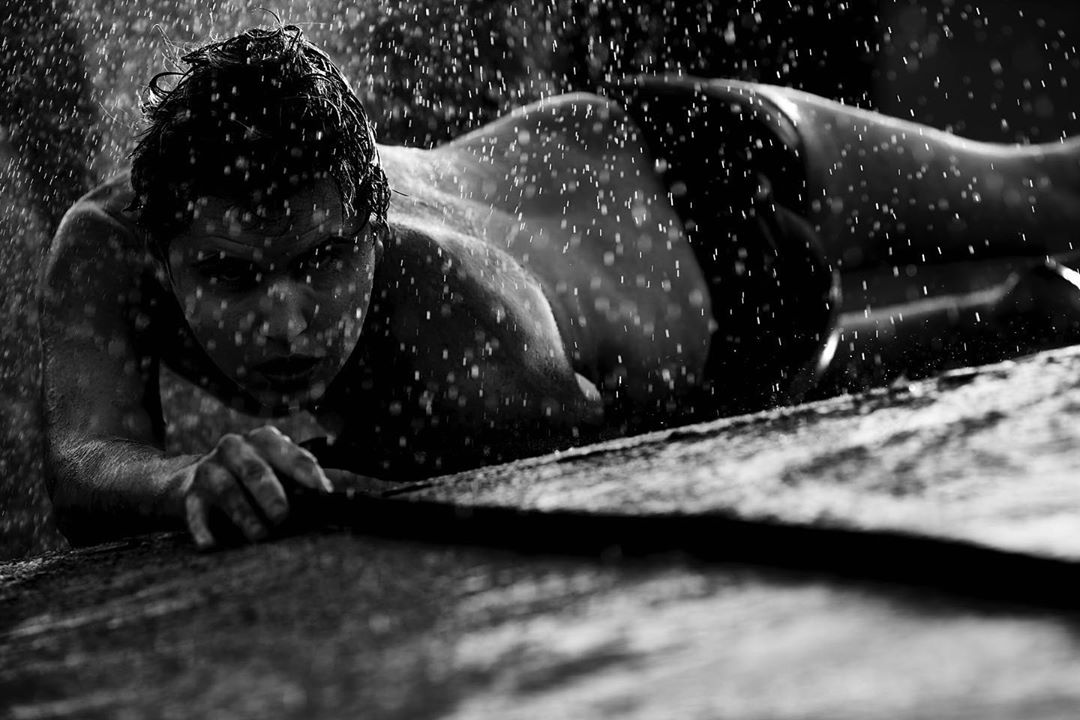 Monochrome close-up of a wet, shirtless man lying on a textured surface, with droplets of water falling on his face and body.